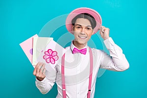 Photo of positive guy wear white shirt bow tie touching pink headwear demonstrate postcards in arm isolated on blue