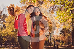 Photo of positive two girls buddies rest relax in autumn park enjoy collect maple leaves hug embrace wear season