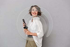 Photo of positive woman 20s holding takeaway coffee and using mo