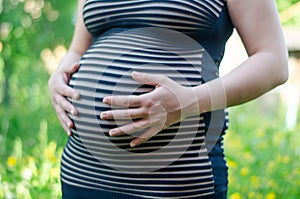 Photo of a pregnant girl in a striped dress with hands on her stomach. Belly close up