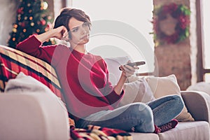 Photo of pretty bored young lady hold remote control hand fist head sitting sofa wear red sweater jeans socks indoors