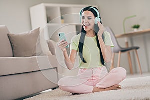 Photo of pretty carefree girl sit carpet crossed legs open mouth hold telephone wear earphones green t-shirt pink