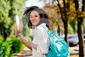 Photo of pretty cheerful lady wear white shirt rucksack walking college chatting modern device outside urban city park