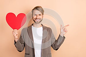 Photo of pretty classy gentleman dressed blazer holding big red heart pointing finger empty space isolated beige color