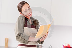 Photo of pretty cute housemaid wear brown apron smiling looking cooking book talking modern device indoors room home