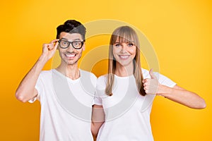 Photo of pretty cute young couple wear white t-shirt spectacles showing thumbs up isolated yellow color background