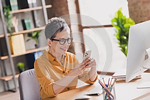 Photo of pretty retired woman hold device sit table dressed yellow formalwear modern workplace working home distance