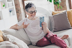 Photo of pretty retired woman hold device video call sofa wear white outfit home flat rest relax spend free spare time