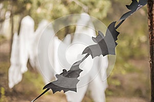 A photo project for Halloween in nature. A garland of black drawn bats against the backdrop of three white ghosts in a forest of g