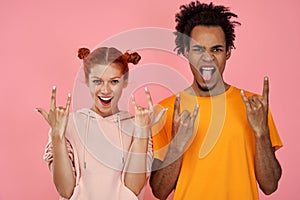 Photo of punky joyful black man and ginger woman hear music of favourite band, show rock n roll gesture, being fan of
