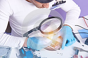 Photo of radioman with soldering iron doing repairs work through magnifying glass, wearing white sweatshirt and blue gloves,