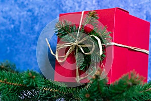 A photo of a red color gift box that stands on a coniferous branches, against a wall with an abstract blue color texture