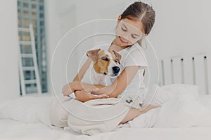 Photo of relaxed beautiful small girl plays with pedigree dog, embraces favourite animal, dressed in casual pyjamas, sits on bed,