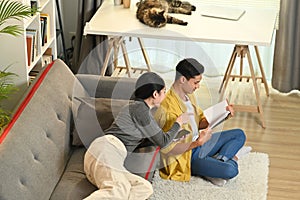 Photo of relaxed married young couple reading book, enjoying leisure time on weekend at home