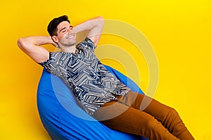 Photo of relaxed sleepy man dressed print shirt brown trousers arms behind head lay on bean bag isolated on yellow color