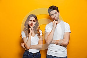 Photo of resented man and woman in casual clothes standing together and touching chin with irritated look, isolated over yellow b