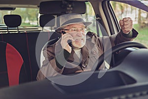 Photo of retired grandfather hold telephone fist hit steering wheel wear brown jacket headwear glasses inside car