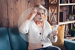Photo of retired woman sit sofa take off specs shocked read news device dressed casual outfit home interior living room