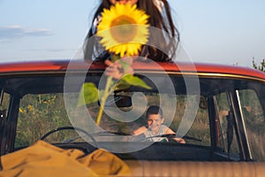 Photo through a retro car windshield, happy teen boy in grey t-shirt leaned on a hood, her sister lies on the roof and