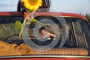 Photo through a retro car windshield, happy teen boy in grey t-shirt leaned on a hood, her sister lies on the roof and