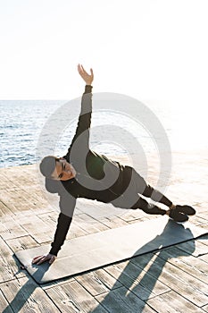 Photo of satisfied sportsman 20s in tracksuit lying on fitness mat and doing plank exercise by seaside in the morning