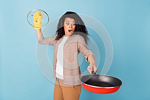 Photo of scared funny dark skin woman dressed checkered shirt holding glove open pan  blue color background