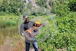 Photo of screaming happy musician against backdrop of summer landscape. young hipster bearded guy holds burning guitar photo