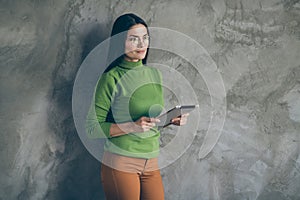 Photo of serious confident pensive woman looking into empty space wearing spectacles holding modern device with hands in