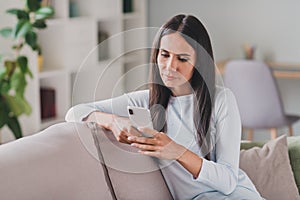 Photo of serious focused pretty brunette young woman hold phone read message sit couch inside home indoors