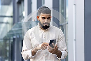 Photo of a serious Indian young man walking down the street near office buildings and using a mobile phone