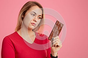 Photo of serious woman holds bar of cocoa chocolate, feels temptation, keeps to diet, dreessed in red outfit, isolated over pink