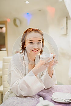 photo of a sexy beautiful reddish woman in white bath gown drinking tea in the fito bar of a spa centre