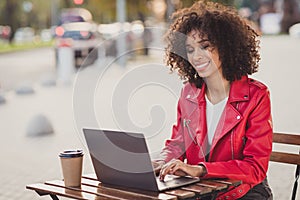 Photo of shiny curly woman dressed red jacked sitting cafe table coffee break typing modern gadget outdoors urban city