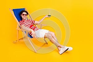 Photo of shiny cute young guy dressed red t-shirt smiling sitting lounge chair drinking cocktail isolated yellow color