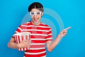 Photo of shocked cute young guy dressed red t-shirt 3d eyewear eating pop corn pointing empty space isolated blue color