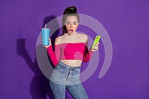 Photo of shocked young lady hold cup cellphone browsing internet wear jeans pink unclothed shoulders top isolated violet