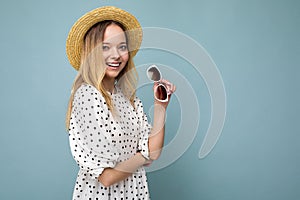 Photo shot of beautiful positive young blonde woman wearing summer casual clothes and holding stylish sunglasses