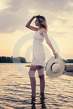 Photo side view of woman with straw hat in her hand. A beautiful young blonde in summer white dress poses against the