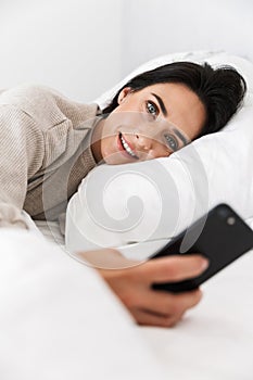 Photo of single woman 30s using smartphone, while lying in bed with white linen at home