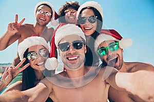 Photo of six multinational friends celebrating new year at tropics taking funny selfies wearing santa claus hats