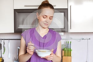 Photo of slim beautiful female model wears purple blouse, holds bowl with cereals, keeps to diet, eats healthy food. Young housewi