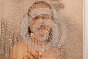 Photo of smiling attractive young woman draws heart on weeping glass shower door, enjoys rest in douche, washing her body,