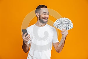 Photo of smiling man 30s in casual wear holding cell phone and fan of money, isolated over yellow background