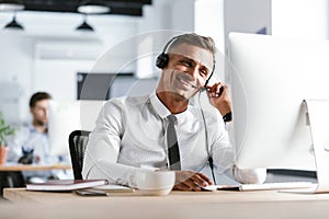 Photo of smiling operator man 30s wearing office clothes and headset, sitting by computer in call center