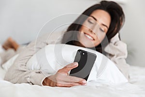 Photo of smiling woman 30s using mobile phone, while lying in bed with white linen in bright room