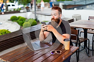 Photo of smiling young bearded man outdoors using laptop computer and mobile phone