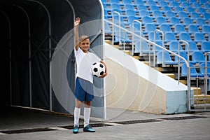 A photo of a sporty kid with a football ball on a stadium background. Outdoors activities. Children training soccer.