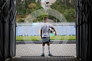 A photo of a sporty kid with a football ball on a stadium background. Outdoors activities. Children training soccer.