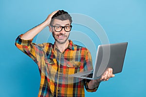 Photo of stressed guilty man in eyeglasses dressed checkered shirt holding laptop hand on head grimace isolated on blue