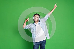 Photo of stubbled young guy raise hands indicate empty space wear headphones denim shirt jeans isolated green color photo